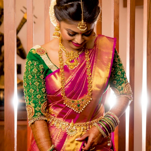An elegant South Indian bride with a pink silk saree and green blouse corrects the pleats in her saree